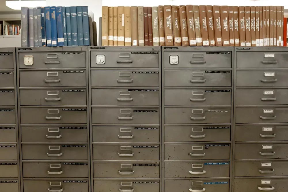Rows of steel filing cabinets topped with decades of bound paper record indexes, from a physical records archive.