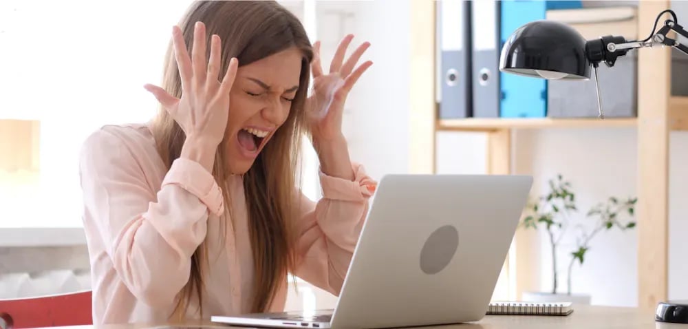 A frustrated woman throws her hands up in front of her laptop in a bright home office.