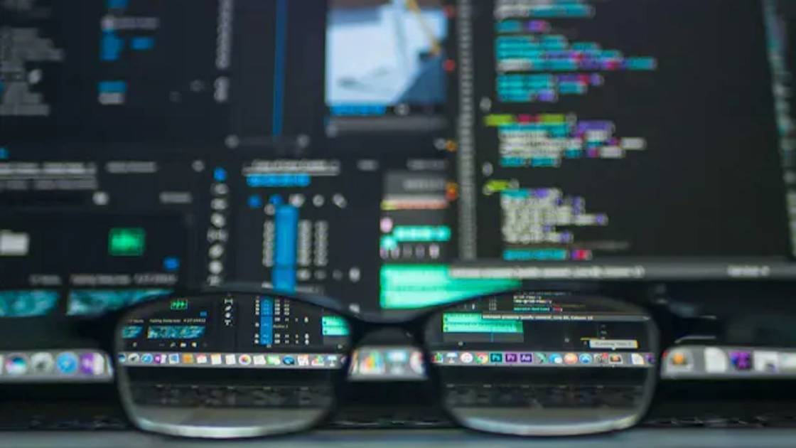 A pair of glasses reflects lines of code and open admin tool windows on a dark monitor.