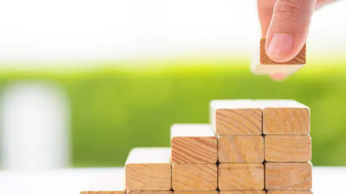 A hand stacking a small wooden block onto a rising staircase of wooden blocks