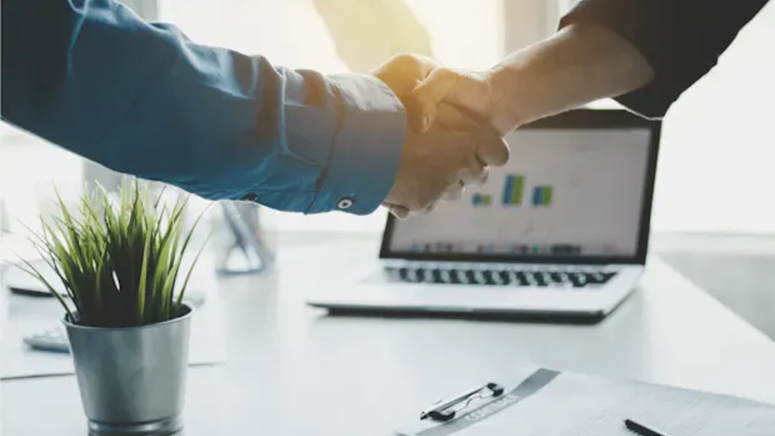 Two colleagues shaking hands over a desk, with a laptop showing a bar chart in the background