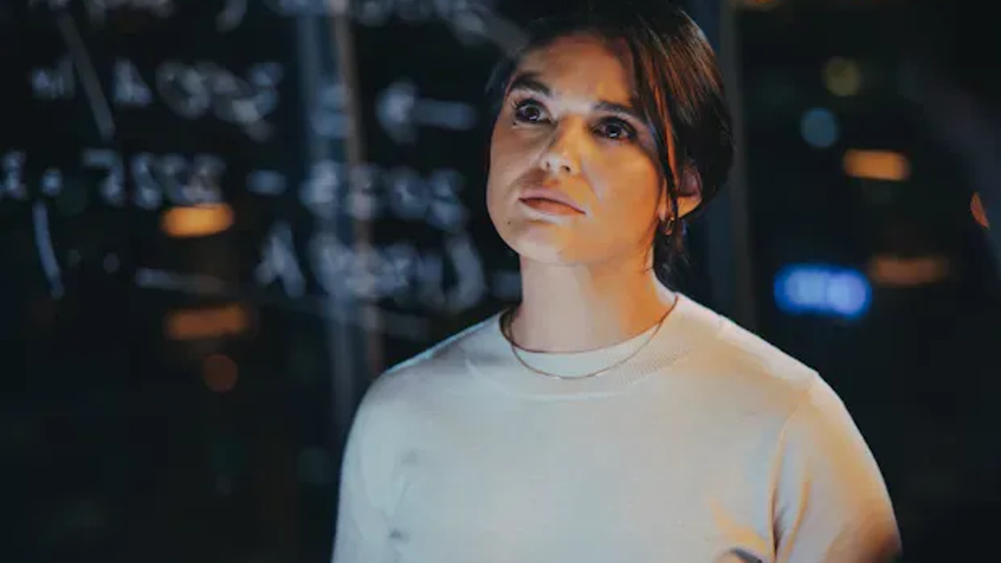 A woman stands in front of a glass wall covered in handwritten equations, lit by warm ambient light, looking upward in thought.