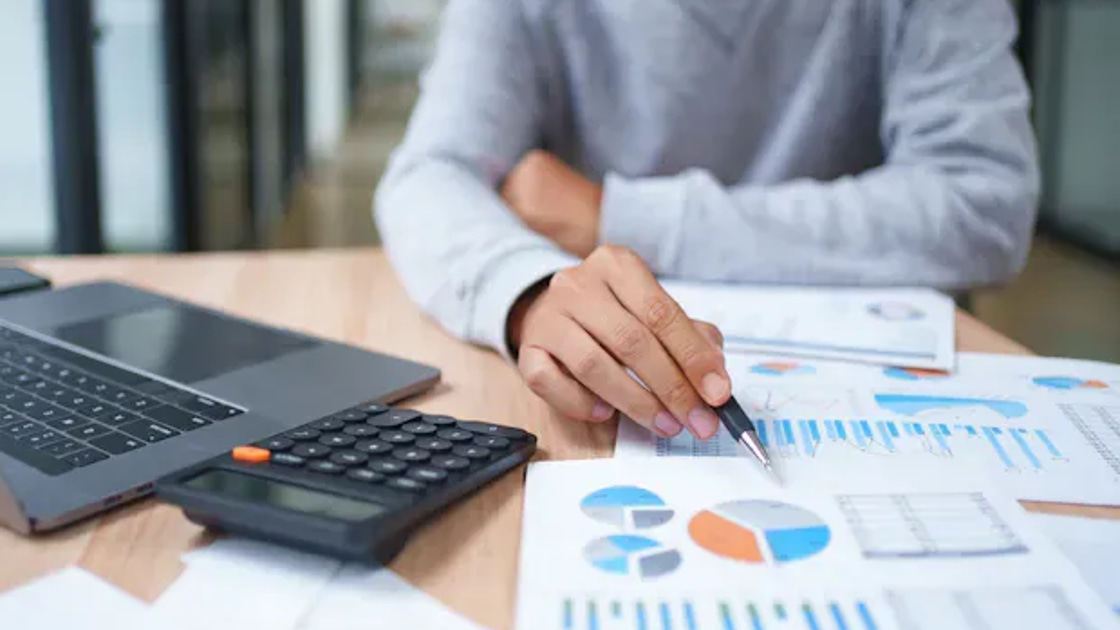 A person reviewing financial charts and budget reports with a calculator and laptop on the desk.