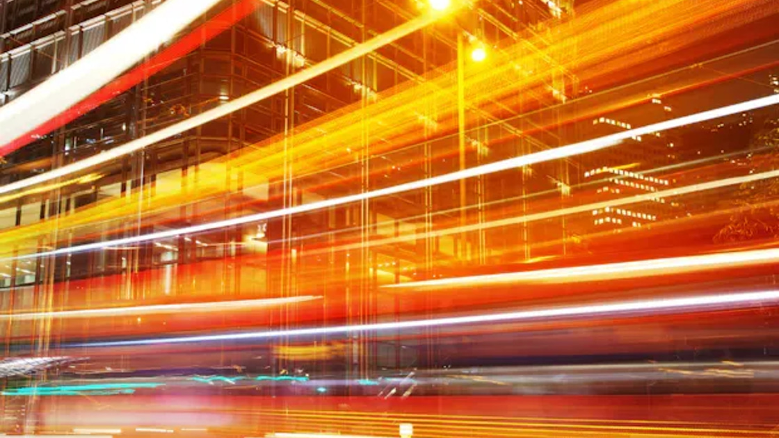 Long-exposure light trails from moving traffic streaking past a building at night, evoking speed and motion.