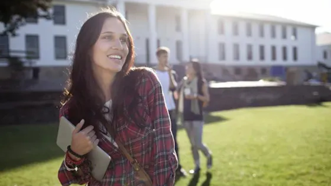 A student carries a tablet across a sunlit college campus lawn while two other students talk in the background in front of a large stone academic building.