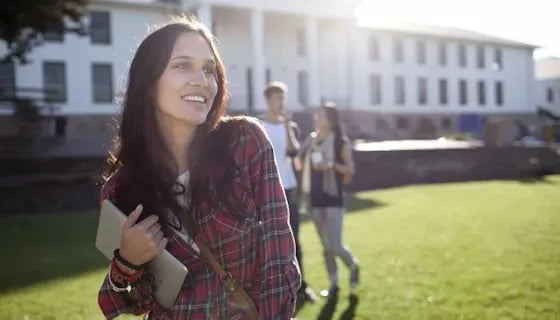 A student carries a tablet across a sunlit college campus lawn while two other students talk in the background in front of a large stone academic building.