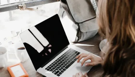 A person typing on a laptop at a wooden desk near a window, with a leather bag and a cup of coffee beside them.