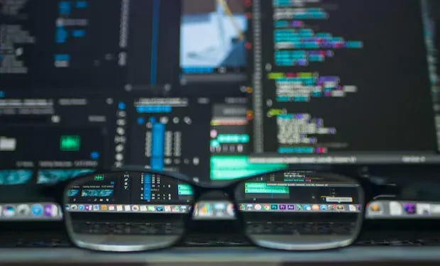 A pair of glasses reflects lines of code and open admin tool windows on a dark monitor.