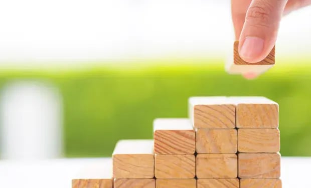 A hand stacking a small wooden block onto a rising staircase of wooden blocks