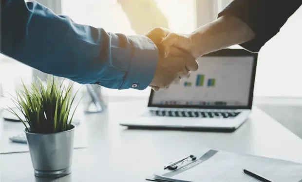 Two colleagues shaking hands over a desk, with a laptop showing a bar chart in the background