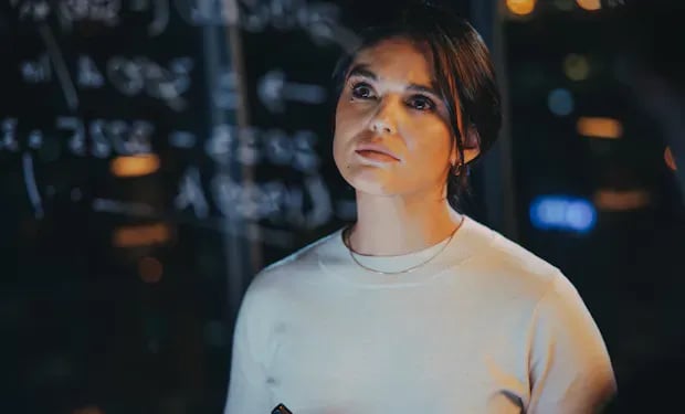 A woman stands in front of a glass wall covered in handwritten equations, lit by warm ambient light, looking upward in thought.