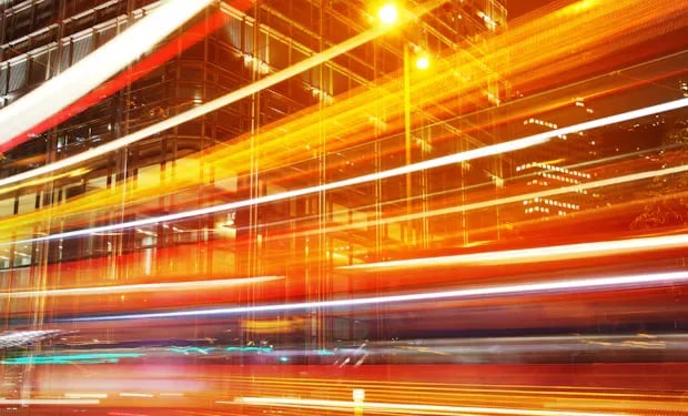 Long-exposure light trails from moving traffic streaking past a building at night, evoking speed and motion.