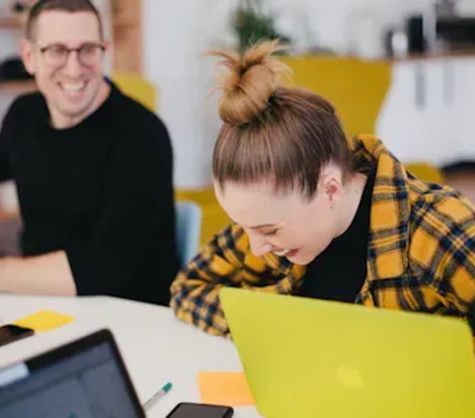 Two colleagues laughing together while working at laptops in an office
